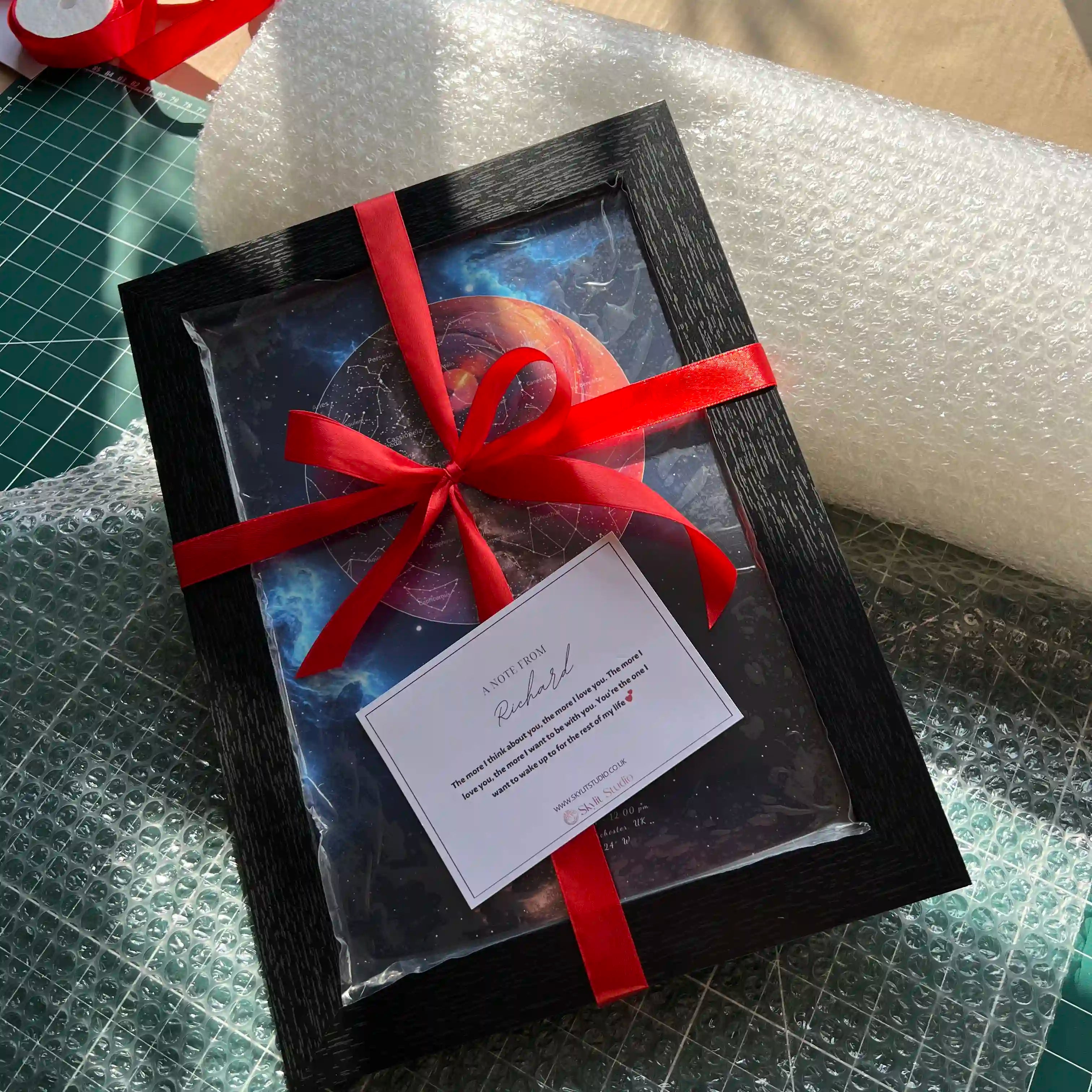 Framed star map gift from Skylit Studio, elegantly wrapped with a red satin ribbon and personalised message note, placed on bubble wrap for secure packaging.