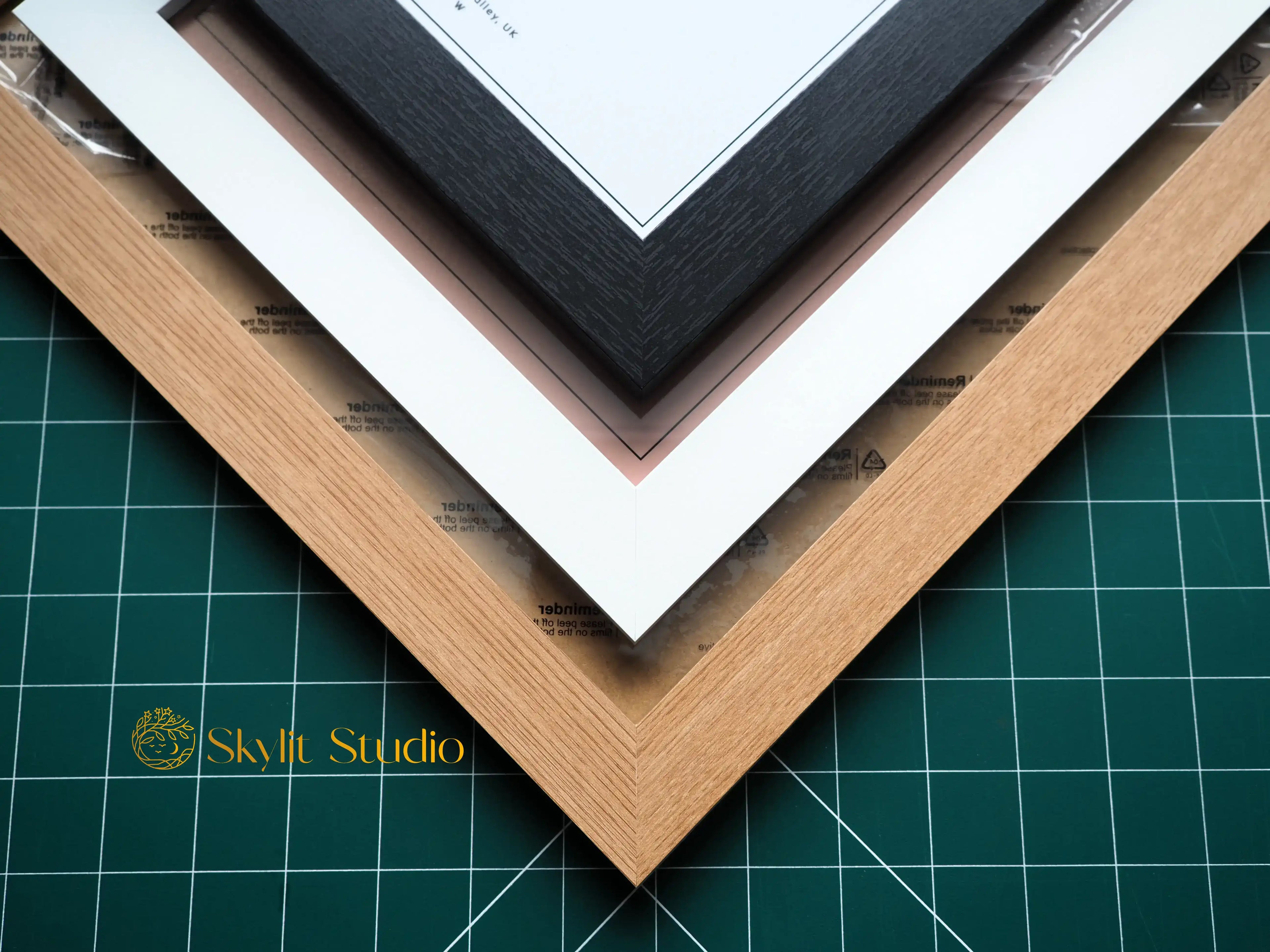 A close-up of three stylish picture frames—black, white, and natural wood—displaying a personalised star map print from Skylit Studio. The frames are arranged in a layered corner view on a green cutting mat, showcasing premium craftsmanship and customisation options for wall art.