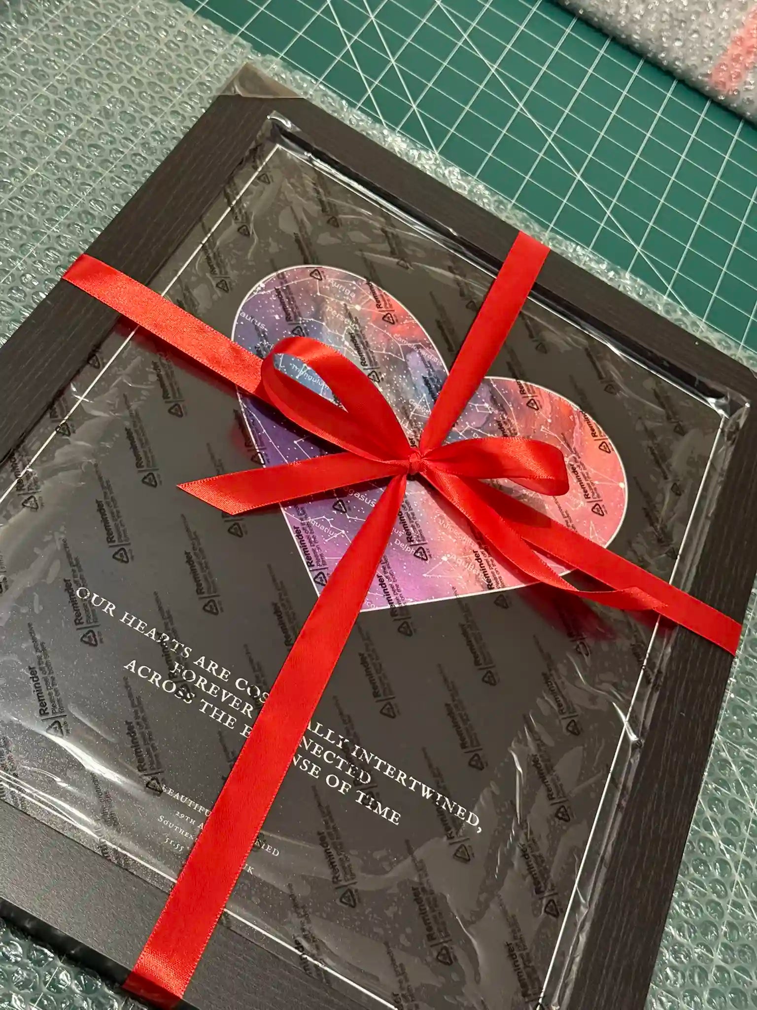 A beautifully wrapped personalised star map print from Skylit Studio, featuring a heart-shaped celestial design on a black background. The framed artwork is secured with protective wrapping and adorned with a vibrant red ribbon, making it a perfect gift for anniversaries, weddings, or special moments.