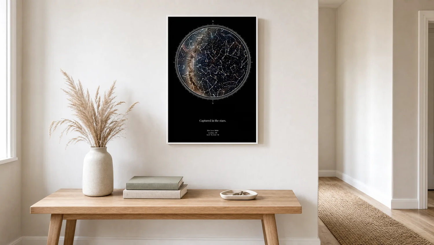 A framed personalised star map print hanging on a pale hallway wall above an oak console table in bright daylight