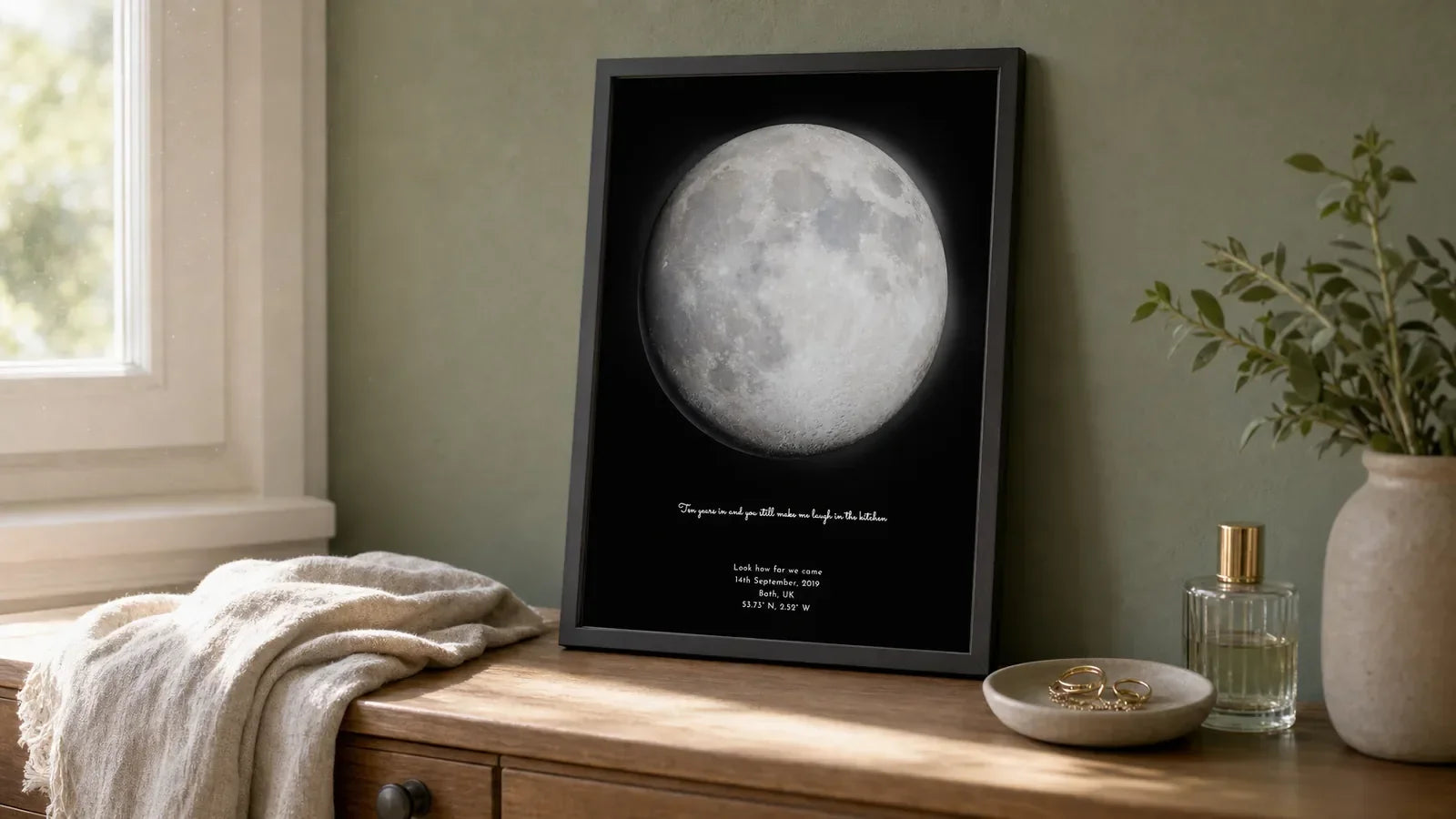 Framed personalised moon phase print leaning on a bedroom dresser in soft late-morning light, beside a ring dish and scent bottle