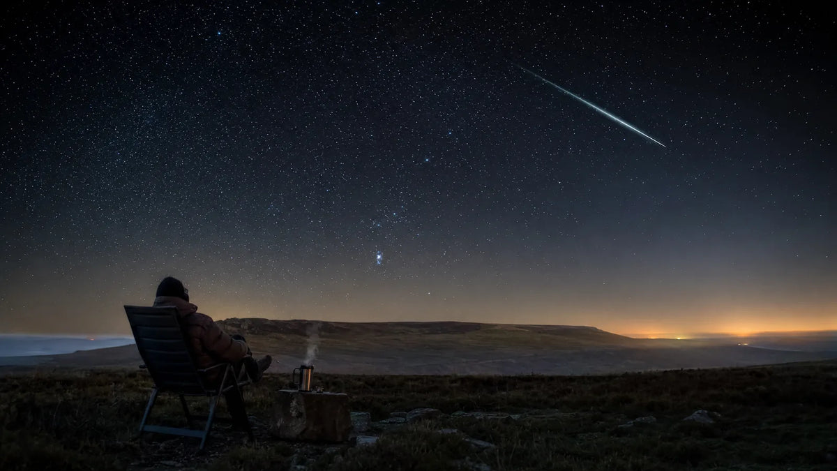A person in a reclining chair on a UK hillside watching a bright meteor cross the sky above Orion before dawn