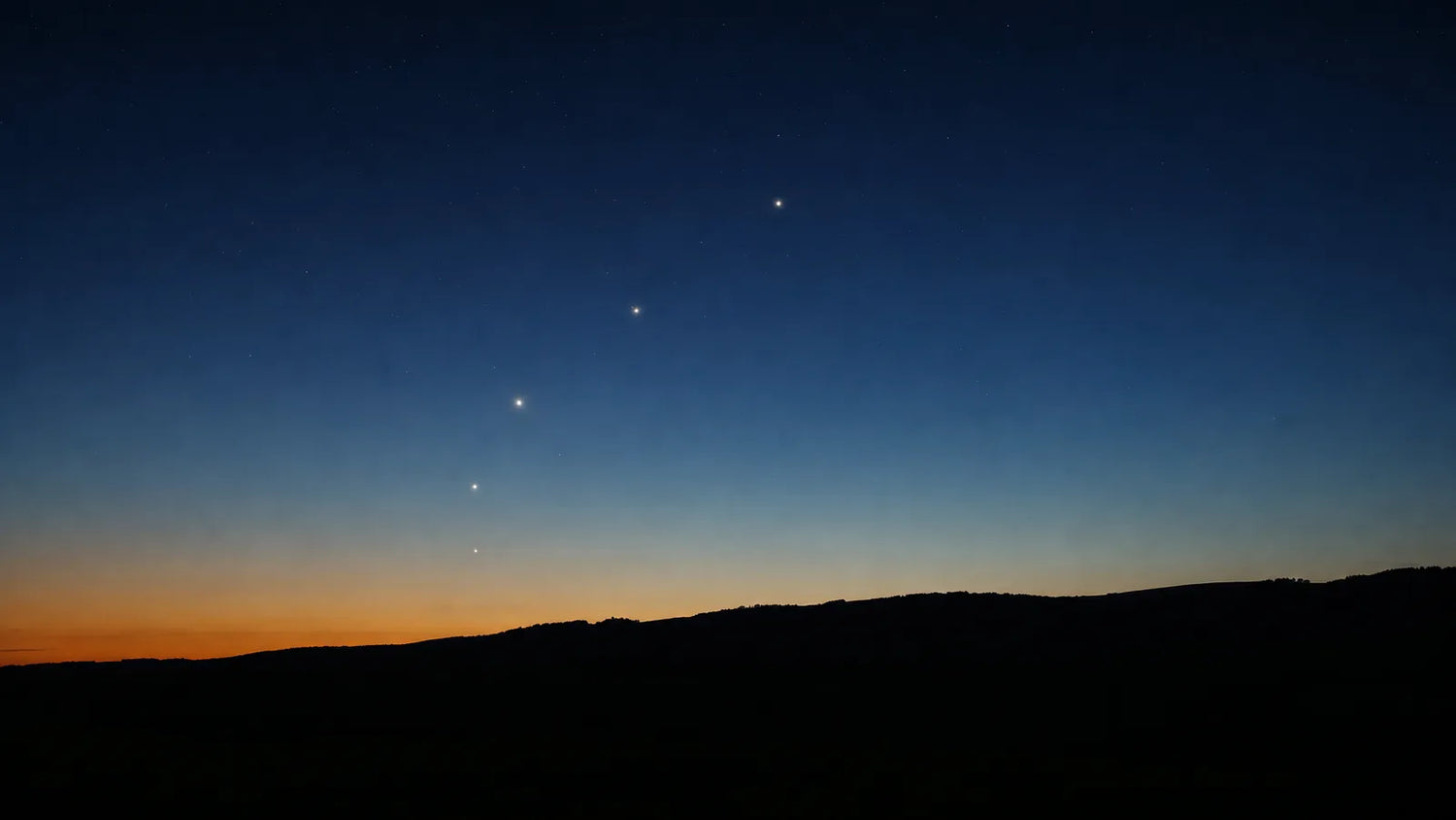 Several bright planets in a loose diagonal line above a dark rural ridgeline in deep blue twilight