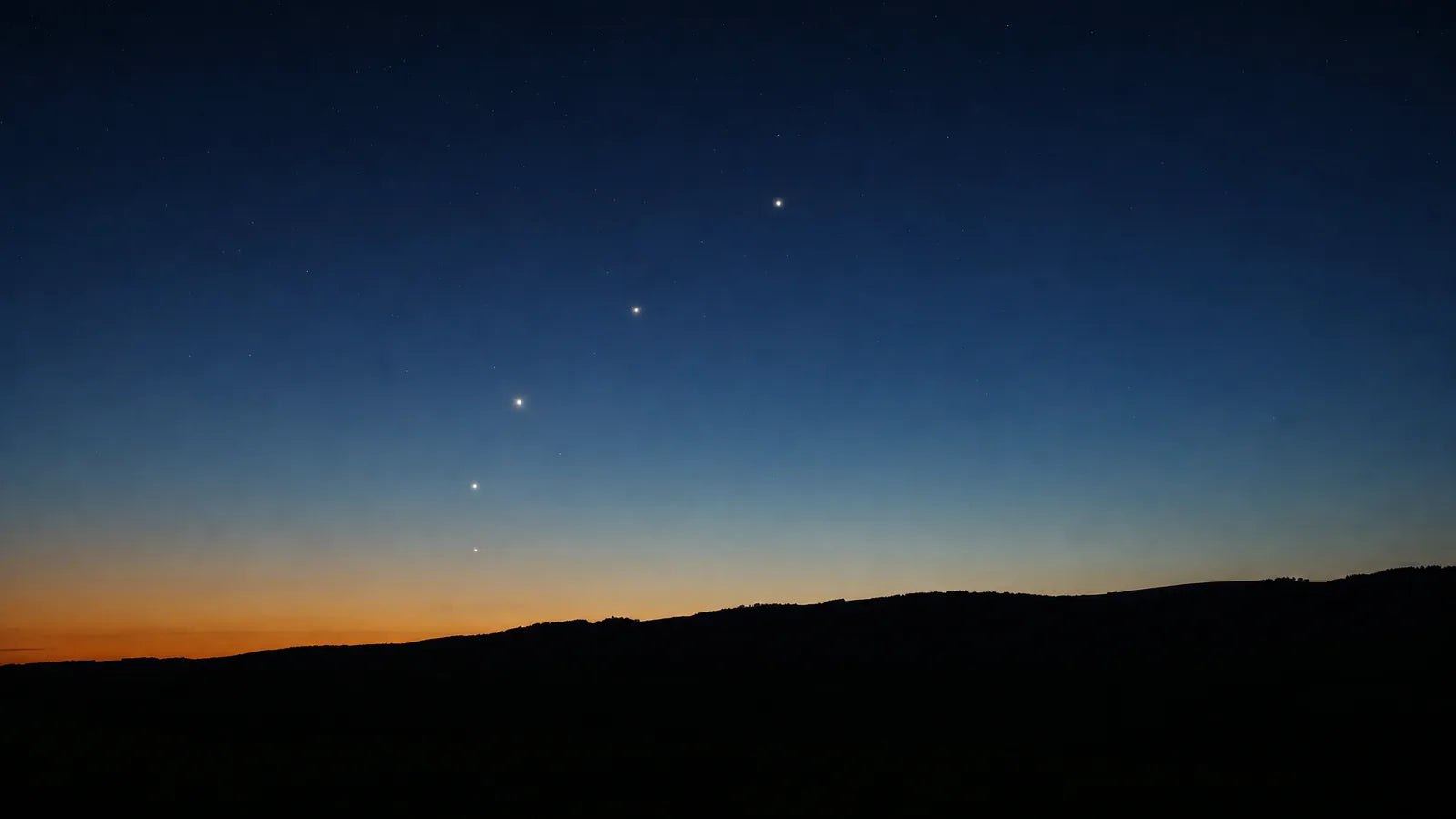 Several bright planets in a loose diagonal line above a dark rural ridgeline in deep blue twilight