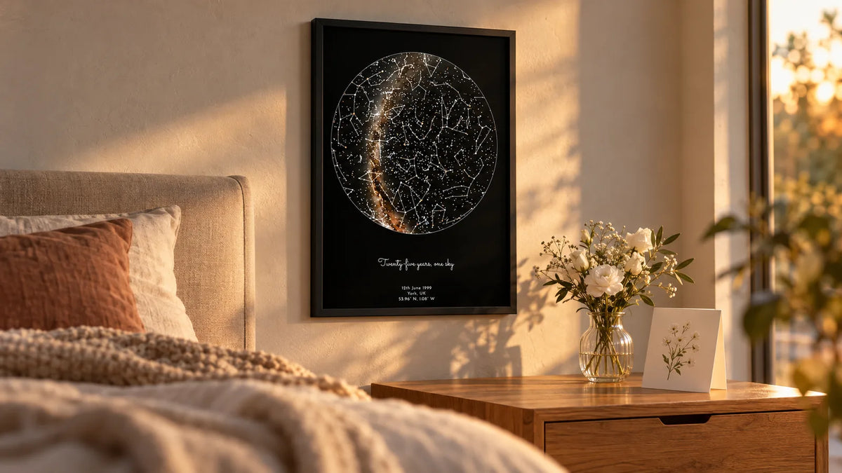 Framed star map print hanging above a bedside table at golden hour, with white flowers and an anniversary card