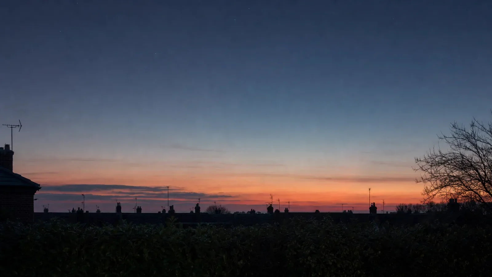 British twilight sky over silhouetted terraced rooftops and chimneys, with the first bright points of light appearing in the deep blue above the amber afterglow