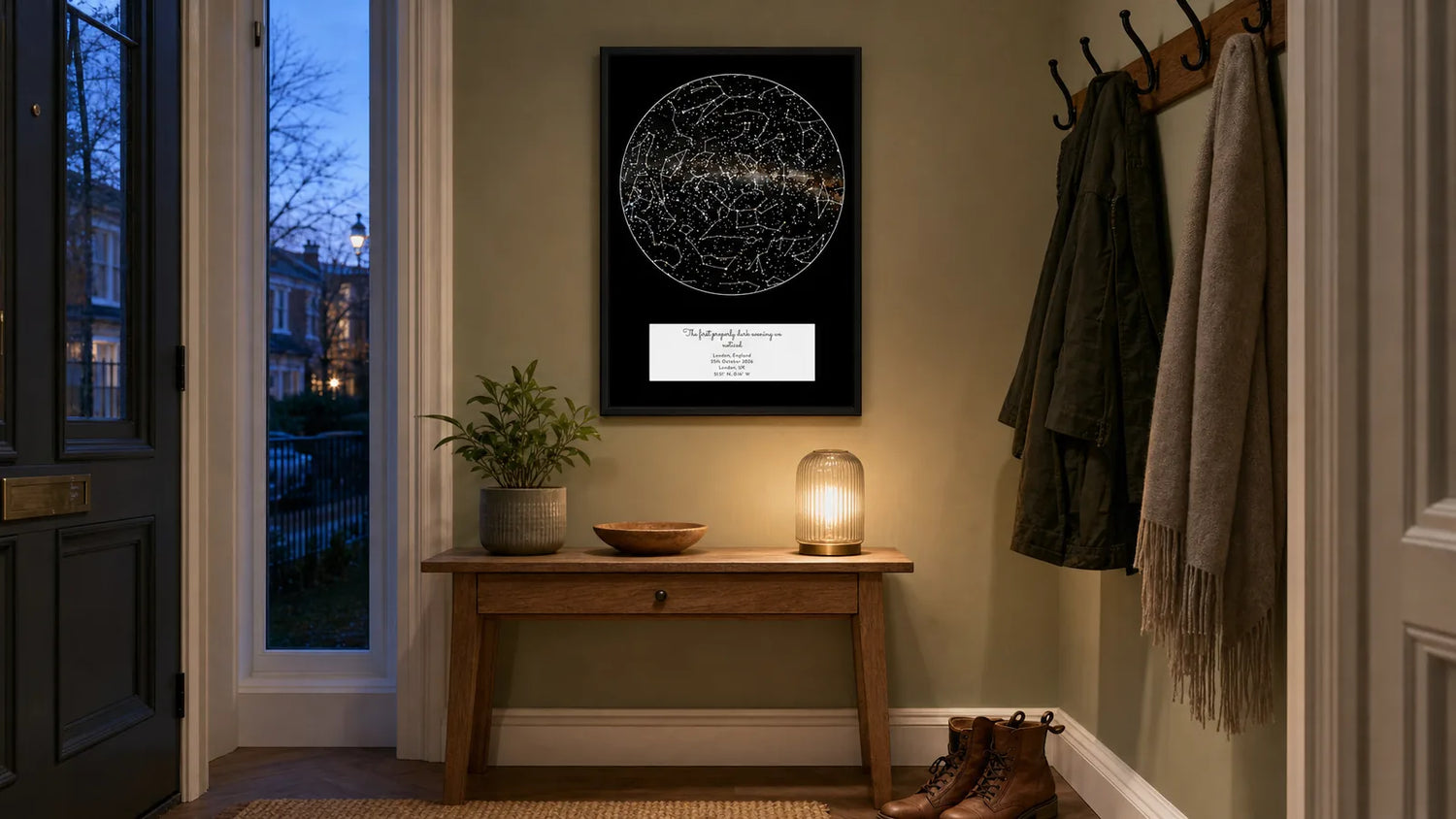 A framed star map print hanging above a console table in a cosy entryway hallway at dusk, blue evening light through the door window beside warm interior lamp light