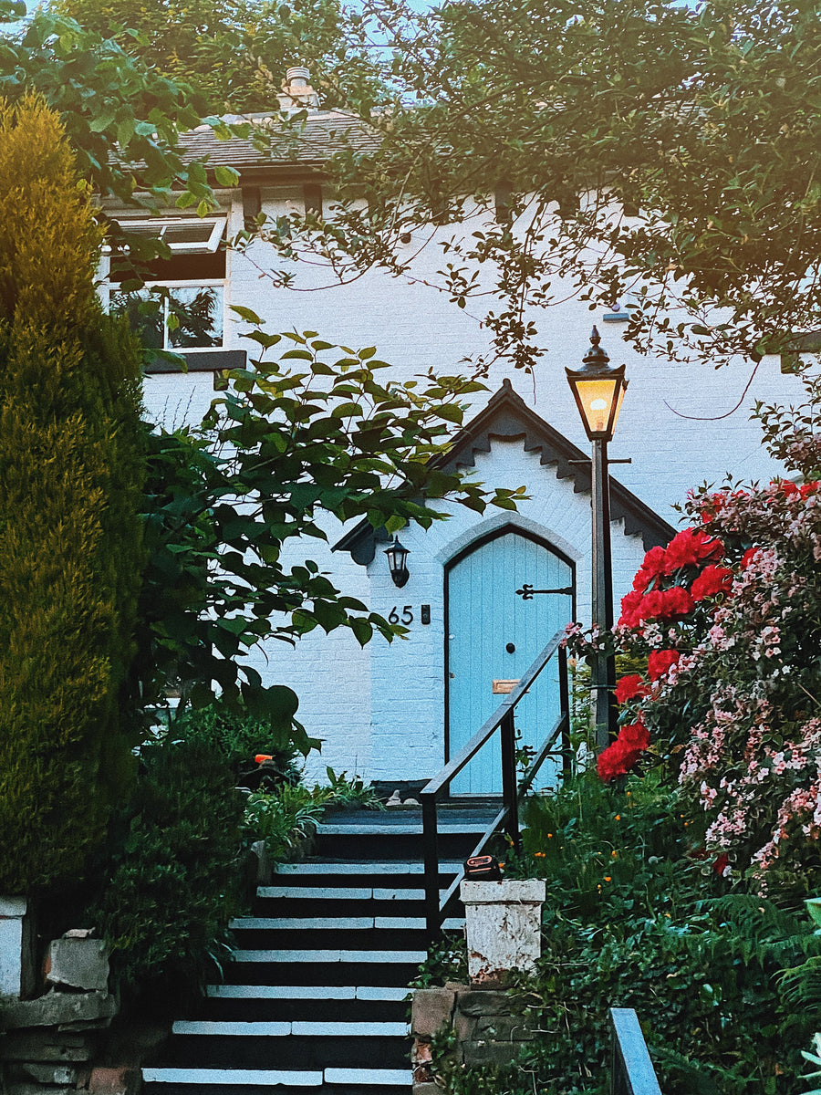 Skylit Studio - Charming cottage entrance with a blue front door, surrounded by lush greenery, stone steps, and vibrant red flowers.