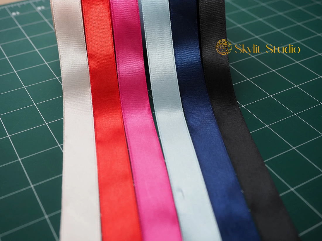 A selection of vibrant satin ribbons in various colours—white, red, pink, light blue, navy blue, and black—laid out on a green cutting mat. Featuring the Skylit Studio logo, these premium ribbons are ideal for gift wrapping, crafts, and personalised packaging.