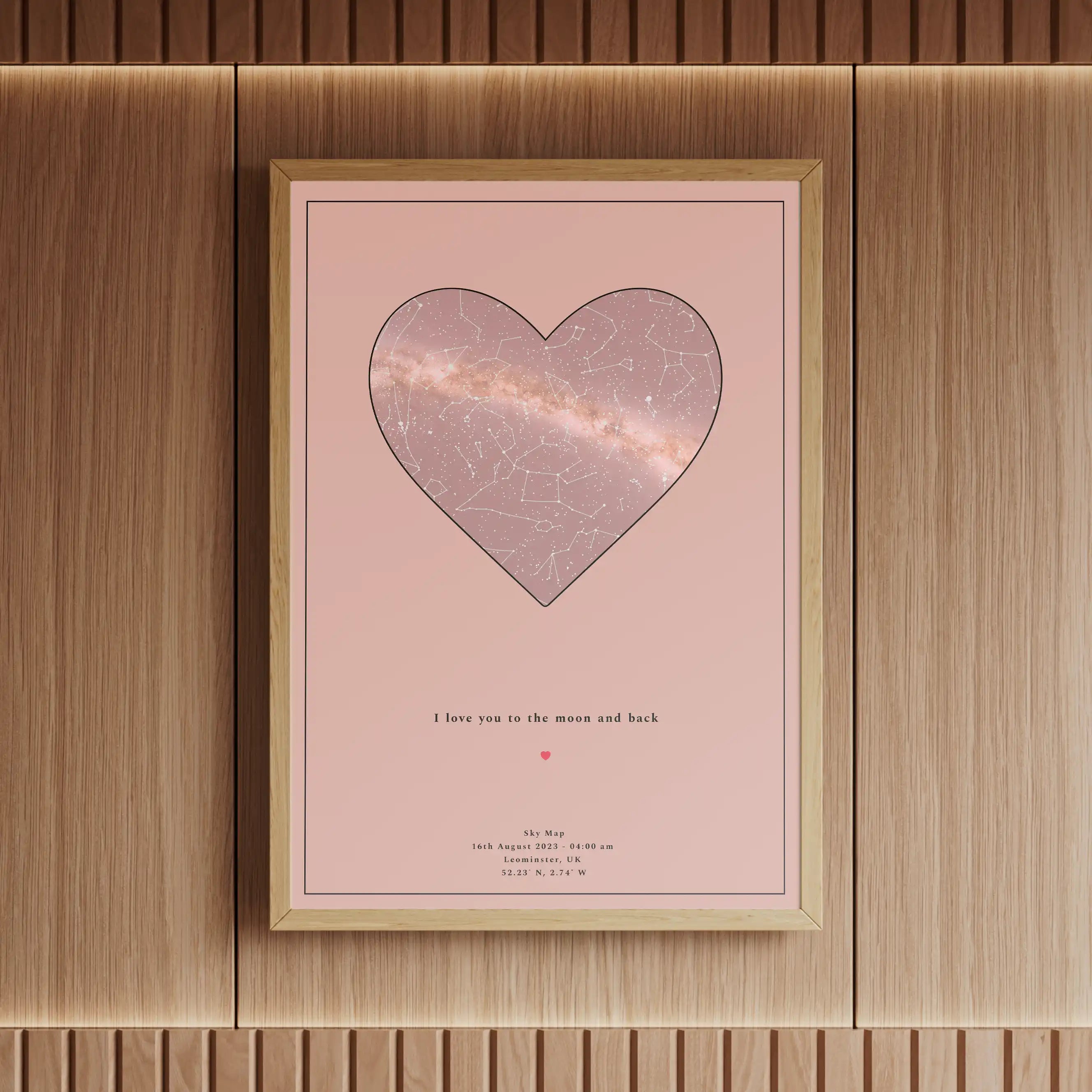 Framed personalised star map print featuring a heart-shaped constellation design on a soft pink background, displayed in an elegant wooden setting with warm lighting.