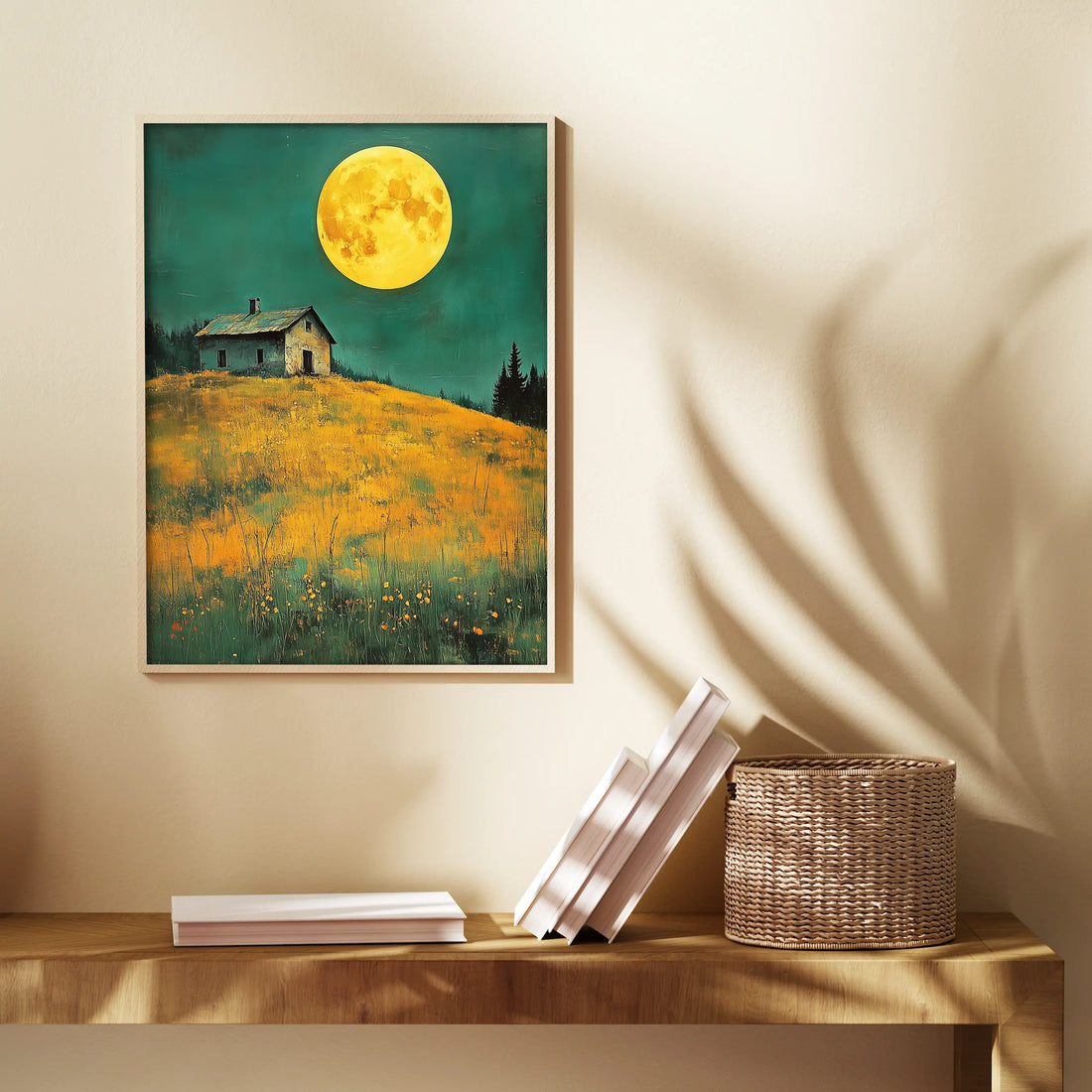 A warm and inviting room corner with a wooden console table displaying a small stack of white books and a woven basket. Above the table, a striking framed artwork features a vibrant yellow moon illuminating a hilltop house surrounded by a field of golden flowers, set against a teal sky. The painting is highlighted by the soft shadows of leaves cast on the wall by ambient lighting.